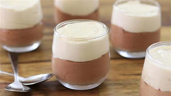 Small glass jars filled with a dessert, consisting of a bottom layer of chocolate mousse and a top layer of vanilla mousse, are arranged on a wooden surface. Two spoons are placed among the jars.