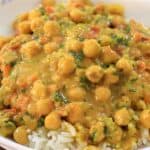 A white bowl with a decorative blue pattern on the rim contains a serving of white rice topped with a chickpea curry. The curry has a golden yellow sauce, mixed with chopped vegetables such as tomatoes and fresh herbs, making for a colorful, appetizing dish.