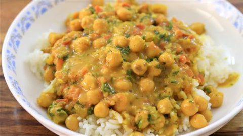 A white bowl with a decorative blue pattern on the rim contains a serving of white rice topped with a chickpea curry. The curry has a golden yellow sauce, mixed with chopped vegetables such as tomatoes and fresh herbs, making for a colorful, appetizing dish.