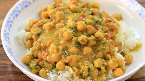 A white bowl with a decorative blue pattern on the rim contains a serving of white rice topped with a chickpea curry. The curry has a golden yellow sauce, mixed with chopped vegetables such as tomatoes and fresh herbs, making for a colorful, appetizing dish.