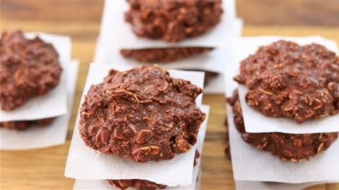 Stacks of chocolate oatmeal cookies are separated by small squares of parchment paper on a wooden surface. The cookies have a rich, chunky texture with visible oats throughout the chocolate dough.