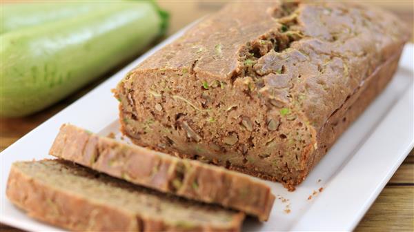 A loaf of freshly baked zucchini bread is displayed on a white rectangular plate. Two slices are cut from the loaf, revealing its moist and textured interior. A whole zucchini is placed in the background on the left side of the image.