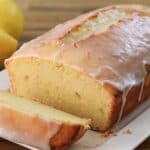 A loaf of lemon pound cake with a slice cut off is placed on a white rectangular plate. The cake has a thick glaze on top and two whole lemons are visible in the background, all set on a wooden surface.