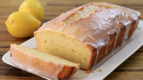 A loaf of lemon pound cake with a slice cut off is placed on a white rectangular plate. The cake has a thick glaze on top and two whole lemons are visible in the background, all set on a wooden surface.