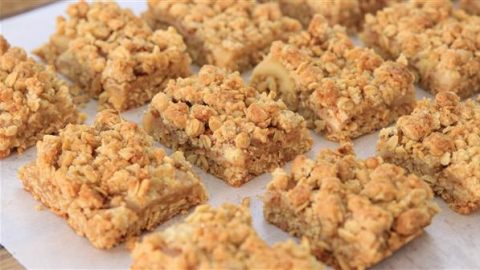 Close-up of a batch of freshly baked dessert bars arranged in rows on parchment paper. The bars have a crumbly oatmeal topping with a golden-brown color, indicating they are likely some kind of fruit crumble or oat streusel bars.