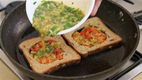 A mixture of eggs, herbs, and vegetables is being poured from a white bowl into the center cut-outs of two slices of bread, which are cooking in a black frying pan. The centers of the bread already contain a mix of diced red, yellow, and green bell peppers.