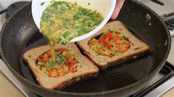 A mixture of eggs, herbs, and vegetables is being poured from a white bowl into the center cut-outs of two slices of bread, which are cooking in a black frying pan. The centers of the bread already contain a mix of diced red, yellow, and green bell peppers.