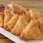 A white rectangular plate holds several golden-brown, triangular samosas neatly arranged in two rows. A small bowl of red dipping sauce sits in the background on the wooden table. The samosas appear crispy and freshly fried.