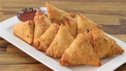 A white rectangular plate holds several golden-brown, triangular samosas neatly arranged in two rows. A small bowl of red dipping sauce sits in the background on the wooden table. The samosas appear crispy and freshly fried.