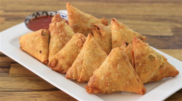 A white rectangular plate holds several golden-brown, triangular samosas neatly arranged in two rows. A small bowl of red dipping sauce sits in the background on the wooden table. The samosas appear crispy and freshly fried.