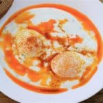 A white plate containing two poached eggs topped with a red paprika sauce and yogurt, served alongside a slice of brown bread. A fork is placed beside the plate on a wooden table with a black and white striped napkin tucked underneath.