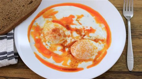 A white plate containing two poached eggs topped with a red paprika sauce and yogurt, served alongside a slice of brown bread. A fork is placed beside the plate on a wooden table with a black and white striped napkin tucked underneath.