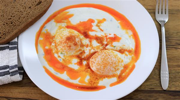 A white plate containing two poached eggs topped with a red paprika sauce and yogurt, served alongside a slice of brown bread. A fork is placed beside the plate on a wooden table with a black and white striped napkin tucked underneath.