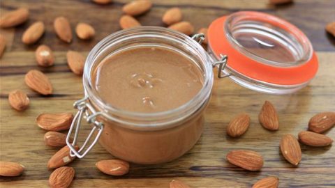 A small glass jar filled with creamy almond butter sits open on a wooden surface, surrounded by whole almonds. The jar has a metal clasp and an orange rubber seal. Some almond butter is visible on the surface inside the jar.