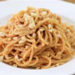 A white plate filled with a serving of spaghetti pasta is topped with a creamy, light brown sauce and sprinkled with small pieces of chopped nuts. The dish is set on a wooden table.