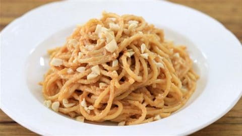 A white plate filled with a serving of spaghetti pasta is topped with a creamy, light brown sauce and sprinkled with small pieces of chopped nuts. The dish is set on a wooden table.