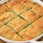 A round white baking dish filled with golden-brown, cut pieces of spanakopita, a Greek dish made with layers of flaky phyllo pastry and a spinach and feta cheese filling. The dish is placed on a wooden surface.