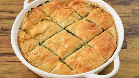 A round white baking dish filled with golden-brown, cut pieces of spanakopita, a Greek dish made with layers of flaky phyllo pastry and a spinach and feta cheese filling. The dish is placed on a wooden surface.