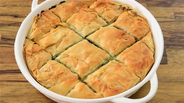 A round white baking dish filled with golden-brown, cut pieces of spanakopita, a Greek dish made with layers of flaky phyllo pastry and a spinach and feta cheese filling. The dish is placed on a wooden surface.