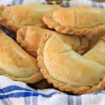 Five baked empanadas in a basket lined with a blue and white checkered cloth. The empanadas have golden-brown crusts and are neatly arranged, showcasing their crimped edges. The background is blurred wood, emphasizing the freshly baked pastries.