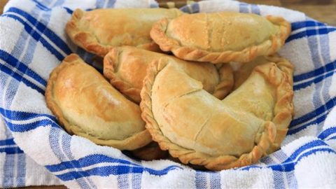 Five baked empanadas in a basket lined with a blue and white checkered cloth. The empanadas have golden-brown crusts and are neatly arranged, showcasing their crimped edges. The background is blurred wood, emphasizing the freshly baked pastries.