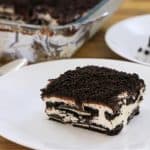 A piece of cookies and cream dessert served on a white plate is in the foreground, featuring layers of crushed chocolate cookies and cream filling. In the background, a baking dish with the remaining dessert and a pair of spoons are visible on a wooden surface.