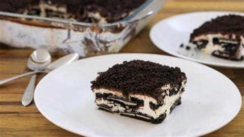 A piece of cookies and cream dessert served on a white plate is in the foreground, featuring layers of crushed chocolate cookies and cream filling. In the background, a baking dish with the remaining dessert and a pair of spoons are visible on a wooden surface.