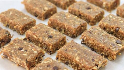 Rows of homemade granola bars, featuring a mixed texture with visible oats, nuts, and chocolate chips, placed on parchment paper. The bars have a natural, rustic appearance, arranged neatly in a grid pattern.