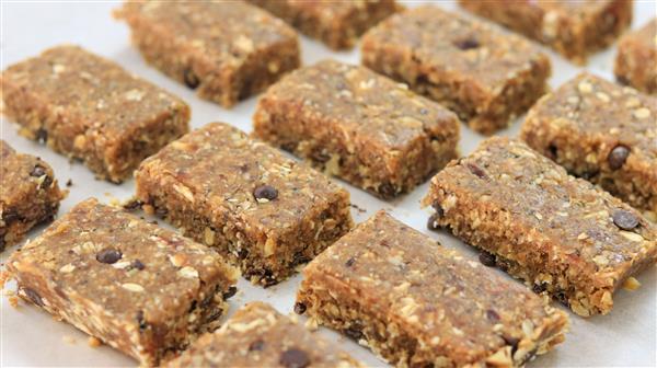 Rows of homemade granola bars, featuring a mixed texture with visible oats, nuts, and chocolate chips, placed on parchment paper. The bars have a natural, rustic appearance, arranged neatly in a grid pattern.