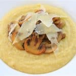 A white bowl filled with creamy yellow polenta, topped with sautéed mushrooms and shavings of Parmesan cheese. The dish is presented on a wooden surface.