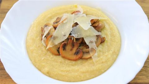 A white bowl filled with creamy yellow polenta, topped with sautéed mushrooms and shavings of Parmesan cheese. The dish is presented on a wooden surface.