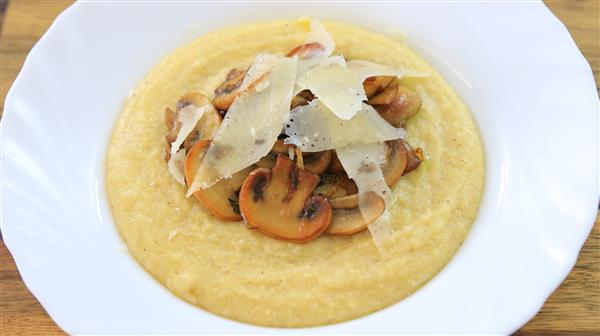 A white bowl filled with creamy yellow polenta, topped with sautéed mushrooms and shavings of Parmesan cheese. The dish is presented on a wooden surface.