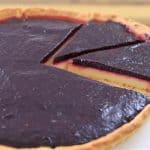 A close-up of a pie with a dark purple filling and a flaky crust. One slice has been partially removed, revealing a yellow custard layer beneath the filling. The pie is placed on a white plate and set on a wooden surface.