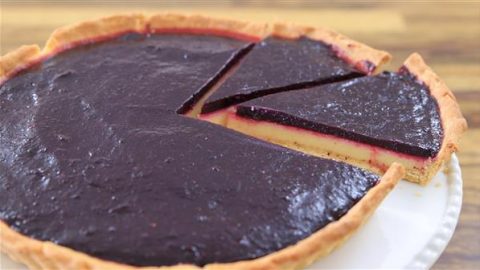 A close-up of a pie with a dark purple filling and a flaky crust. One slice has been partially removed, revealing a yellow custard layer beneath the filling. The pie is placed on a white plate and set on a wooden surface.