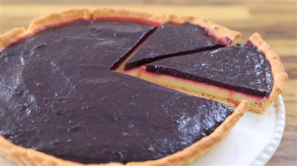 A close-up of a pie with a dark purple filling and a flaky crust. One slice has been partially removed, revealing a yellow custard layer beneath the filling. The pie is placed on a white plate and set on a wooden surface.