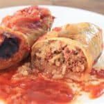 A close-up of two stuffed cabbage rolls on a white plate. One roll is cut in half, revealing a filling of rice, ground meat, and spices. Both rolls are topped with a layer of tomato sauce. The background shows a wooden surface.