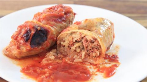 A close-up of two stuffed cabbage rolls on a white plate. One roll is cut in half, revealing a filling of rice, ground meat, and spices. Both rolls are topped with a layer of tomato sauce. The background shows a wooden surface.