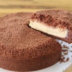 A hand holding a cake server lifts a slice from a round, chocolate crumb-covered cake on a white plate. The cake has a thick layer of creamy filling sandwiched between the chocolate crumb layers, and the background features a wooden table.