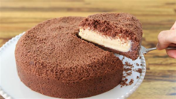 A hand holding a cake server lifts a slice from a round, chocolate crumb-covered cake on a white plate. The cake has a thick layer of creamy filling sandwiched between the chocolate crumb layers, and the background features a wooden table.