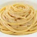 A white plate contains a neatly coiled serving of thick spaghetti noodles, lightly coated in a creamy sauce and sprinkled with ground black pepper. The pasta appears appetizing and is arranged in a spiral pattern.