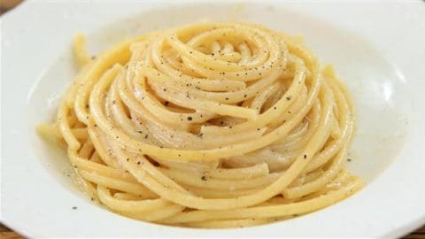 A white plate contains a neatly coiled serving of thick spaghetti noodles, lightly coated in a creamy sauce and sprinkled with ground black pepper. The pasta appears appetizing and is arranged in a spiral pattern.