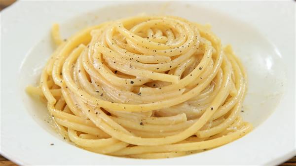 A white plate contains a neatly coiled serving of thick spaghetti noodles, lightly coated in a creamy sauce and sprinkled with ground black pepper. The pasta appears appetizing and is arranged in a spiral pattern.