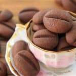 A teacup filled with several chocolate coffee bean-shaped cookies sits on a matching saucer. More cookies are scattered on the wooden table around the cup. The cookies are dark brown with a central line, resembling coffee beans.
