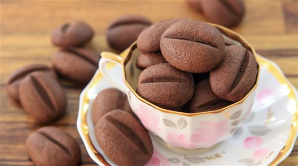 A teacup filled with several chocolate coffee bean-shaped cookies sits on a matching saucer. More cookies are scattered on the wooden table around the cup. The cookies are dark brown with a central line, resembling coffee beans.