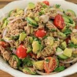 A large, white bowl filled with a colorful salad. The salad has chunks of tuna, sliced red onions, halved cherry tomatoes, diced avocados, cucumber slices, and fresh cilantro. The bowl is placed on a wooden surface.