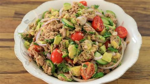 A large, white bowl filled with a colorful salad. The salad has chunks of tuna, sliced red onions, halved cherry tomatoes, diced avocados, cucumber slices, and fresh cilantro. The bowl is placed on a wooden surface.