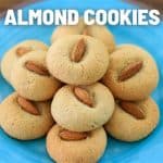 Plate of almond cookies on a bright blue dish, each cookie topped with a whole almond. Text at the top of the image reads "ALMOND COOKIES." The plate rests on a wooden surface.