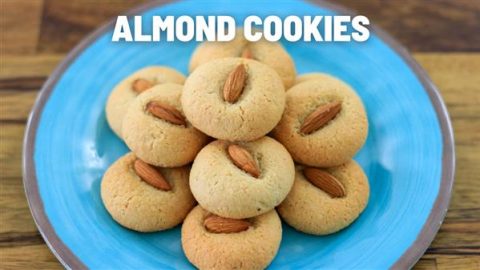 Plate of almond cookies on a bright blue dish, each cookie topped with a whole almond. Text at the top of the image reads "ALMOND COOKIES." The plate rests on a wooden surface.