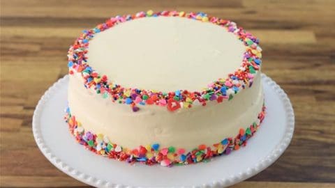 A round, white frosted cake is decorated with colorful sprinkles around the top edge and base. The cake is placed on a white plate, set on a wooden surface.