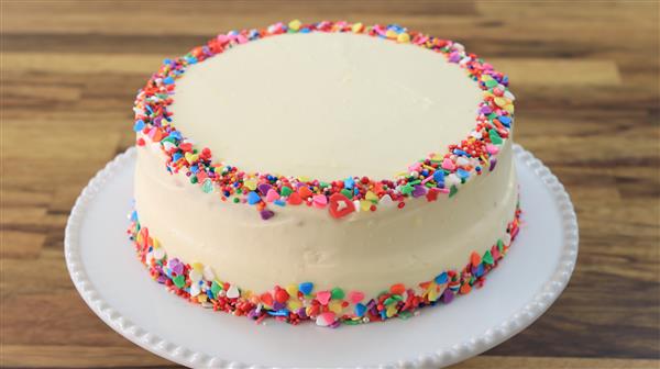 A round, white frosted cake is decorated with colorful sprinkles around the top edge and base. The cake is placed on a white plate, set on a wooden surface.